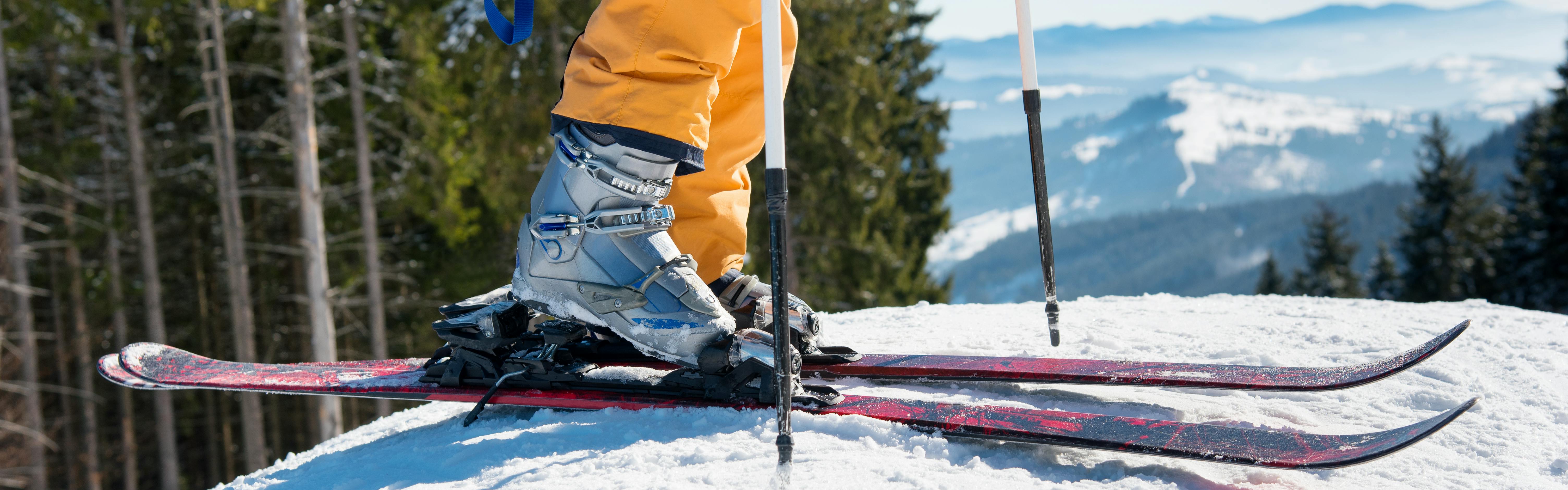 A skier clicking their boot into their ski bindings as they stand on top of a snowy mountain. 