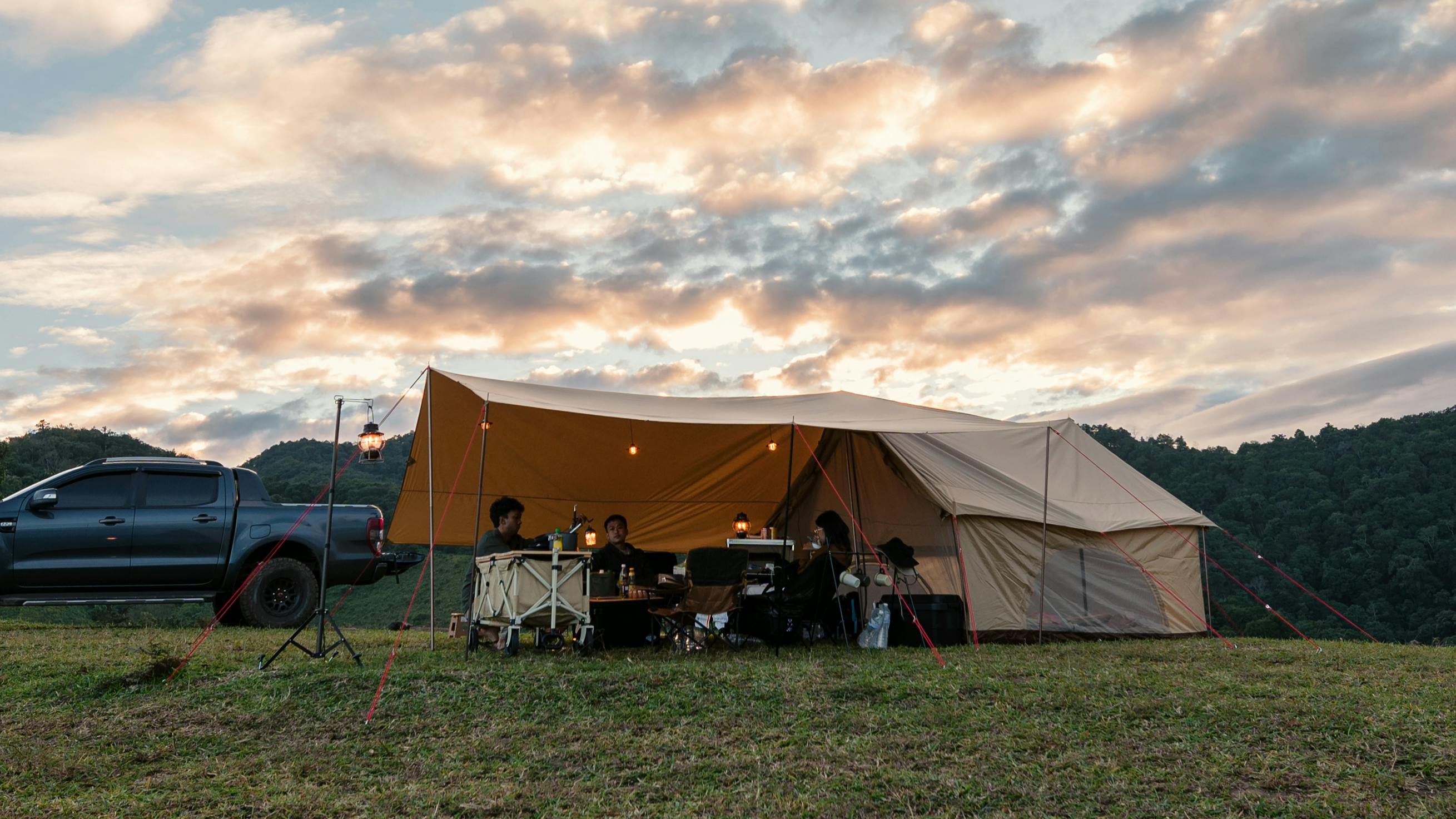 Several campers sit under a shelter outside their tent. A truck is parked next to them and the sun is setting